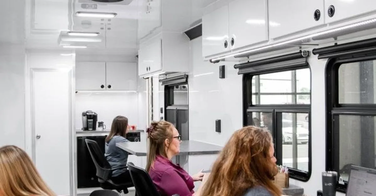 Modern mobile office trailer with workstations, overhead storage, climate control, and staff working comfortably on laptops.