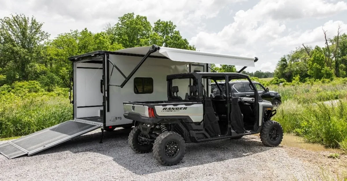 Toy hauler trailer parked outdoors with a side-by-side UTV, featuring a rear loading ramp, awning, and adventure-ready design.