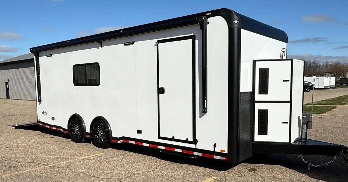 A white enclosed trailer, featuring a side entry door, tinted window, and an exterior awning, parked on a paved lot.