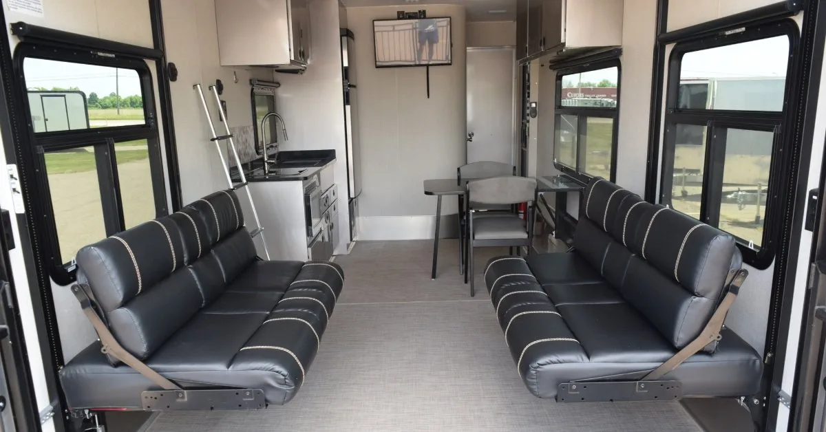 The interior of a trailer, featuring a living space with two sofa-style benches, windows on both sides, overhead storage cabinets, a compact kitchenette, a small dining table with chairs, and a wall-mounted TV.