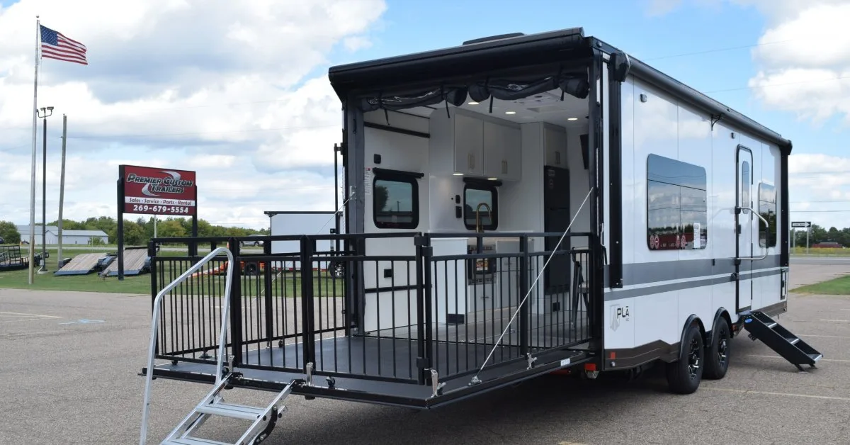 A white trailer parked in a lot, with a fold-down rear patio deck enclosed by black metal railings. The open rear door reveals a living-space interior with cabinetry and seating.