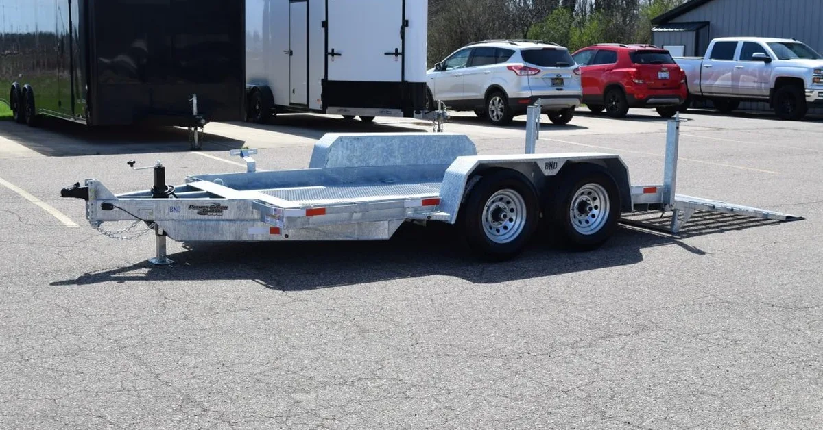 A galvanized steel scissor lift trailer sitting in the parking lot at the trailer company. There are vehicles behind it.